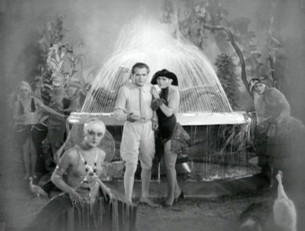 A gaudily dressed group in a garden; there is a fountain and a peacock