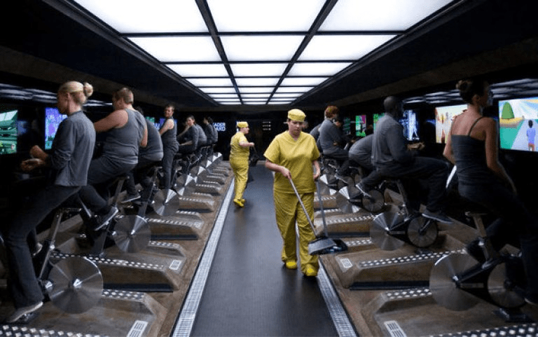 Rows of cyclists on stationary bikes, all staring at screens. In the background, a leaderboard can be glimpsed. Two cleaners in yellow uniforms move down the central aisle